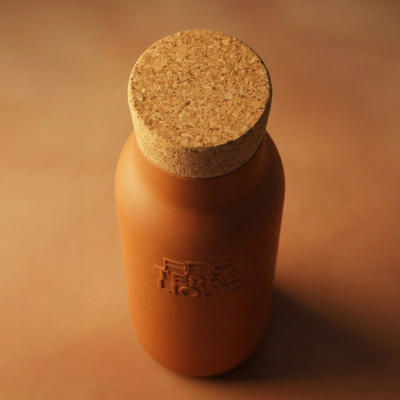 A matte terracotta-colored bottle with a cork stopper, featuring an embossed logo on the front, set against a matching warm brown background.